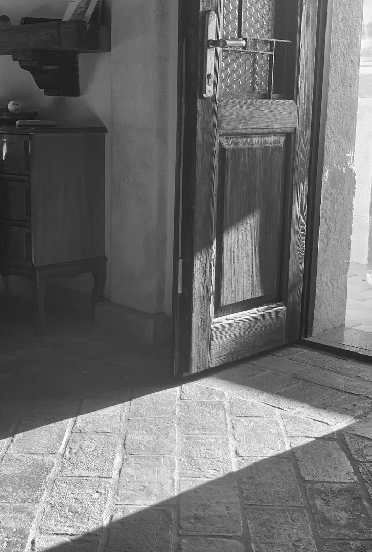 Schwarz-Weiß-Bild einer geöffneten, rustikalen Holztüre, Sonnenlicht strahlt auf den rustikalen Steinboden